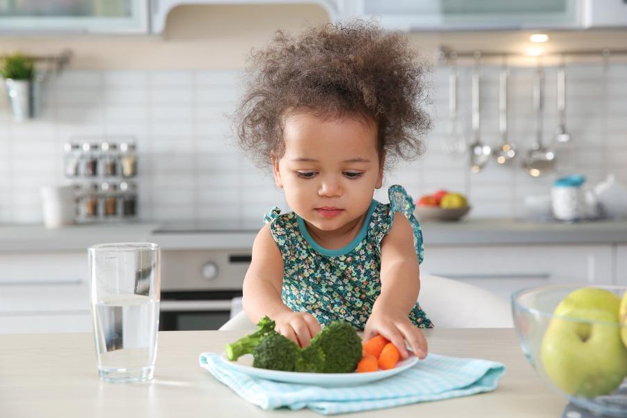 Cute little girl eating vegetables at table in kitchen