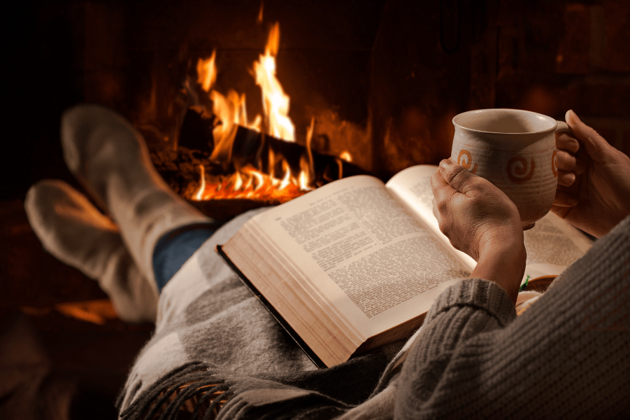 Woman resting with cup of hot drink and book near fireplace