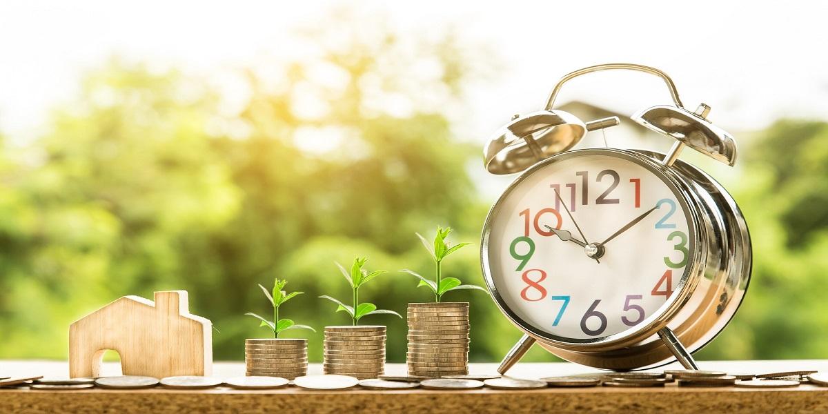 A clock timer, a wooden house with money stacked up with a flower on top