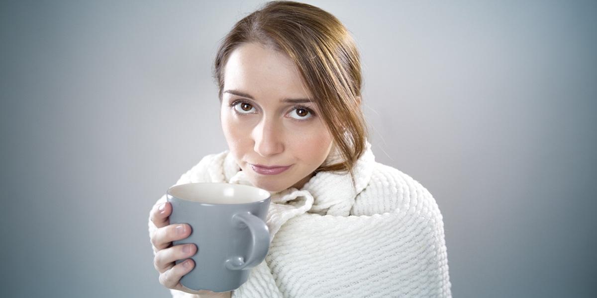 Sick lady in a blanket and holding a hot mug of something.