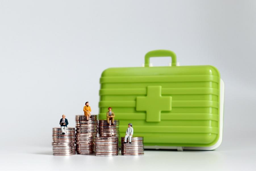 A stack of money with small people sat on and a first aid box in the background